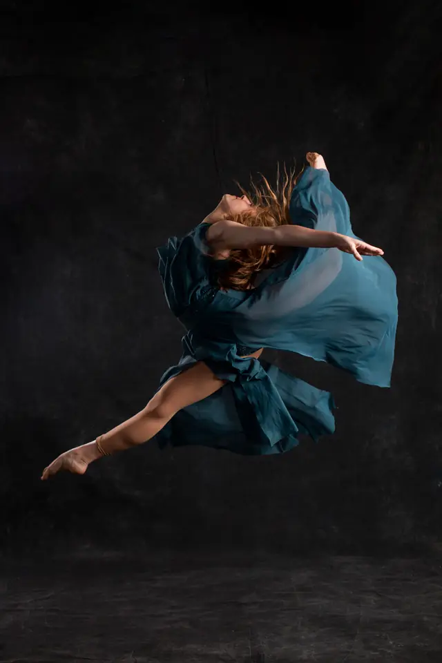 A dancer in a teal dress mid-leap against a dark backdrop