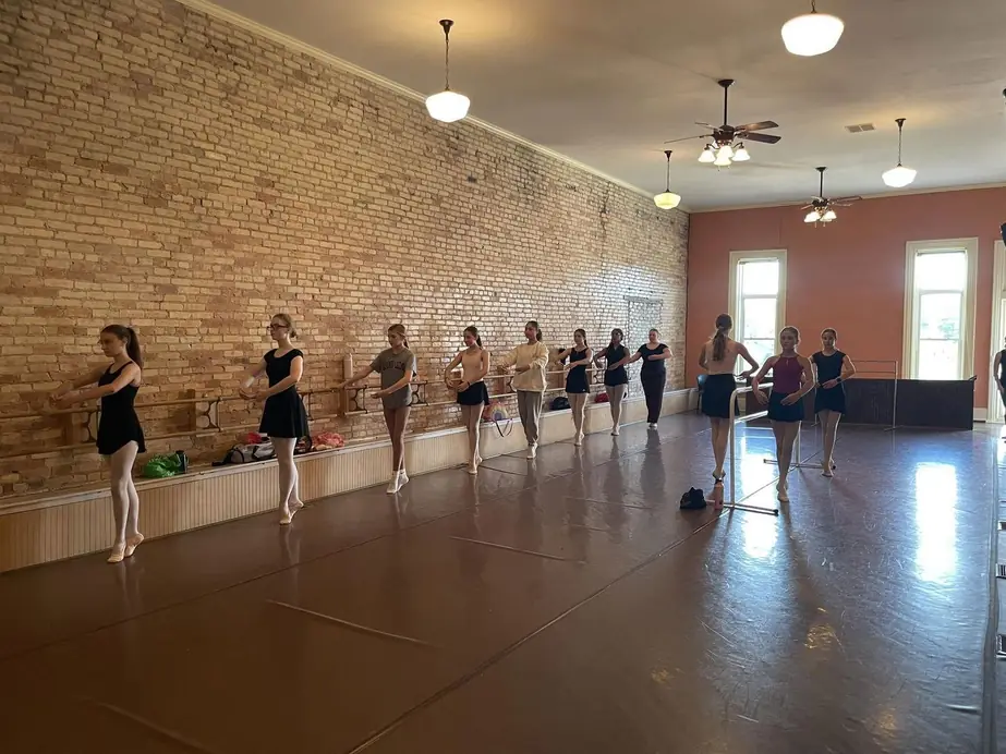 A long line of dancers at the barre along the brick wall