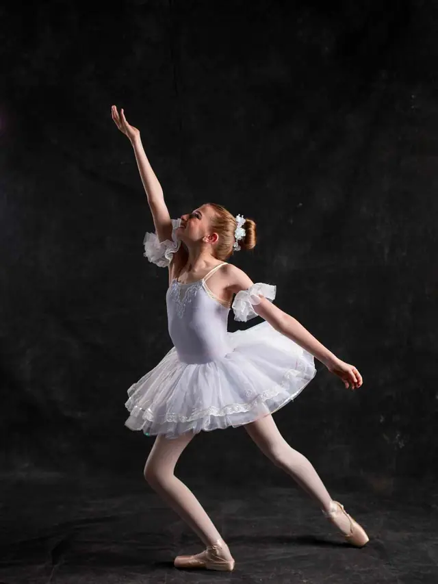 A young ballerina in a white tutu reaching upward against a dark backdrop