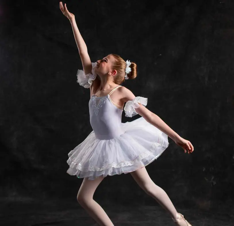 A young ballerina in a white tutu holding a pose against a dark backdrop